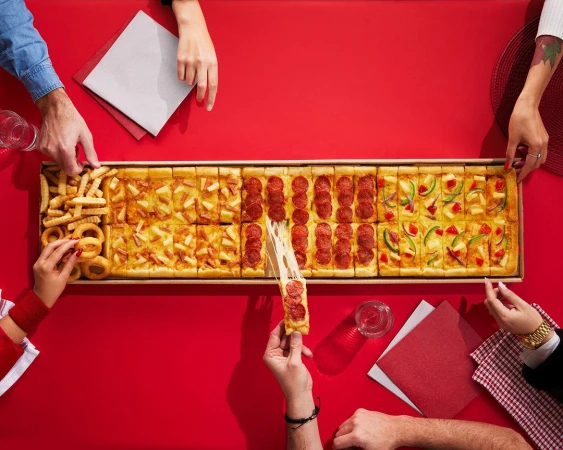 Overhead shot of a long rectangular pizza in a box with various toppings, surrounded by six people's hands on a red table.