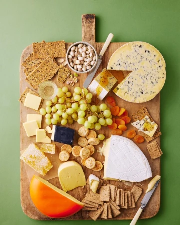 Overhead shot showcases a wooden board topped with cheese, grapes, crackers, nuts, and apricots against a green background.