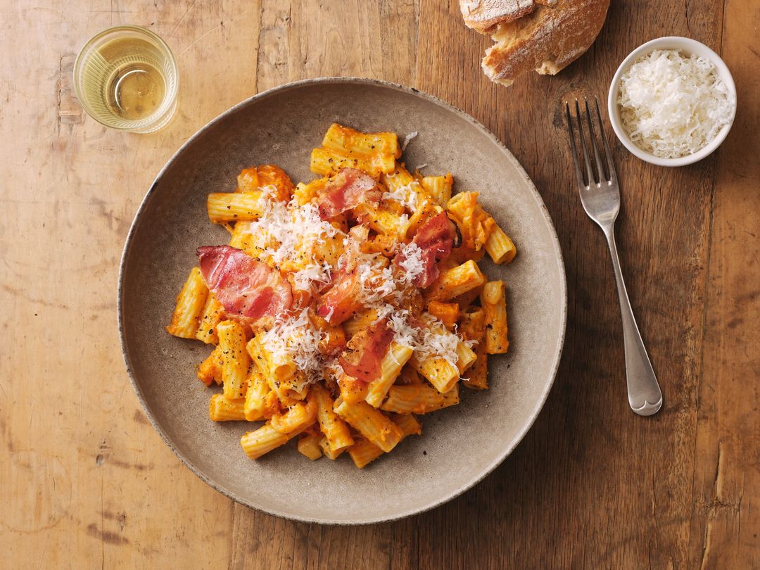 Overhead shot of rigatoni pasta with tomato sauce, bacon, and parmesan cheese on a plate with a glass of white wine and bread.