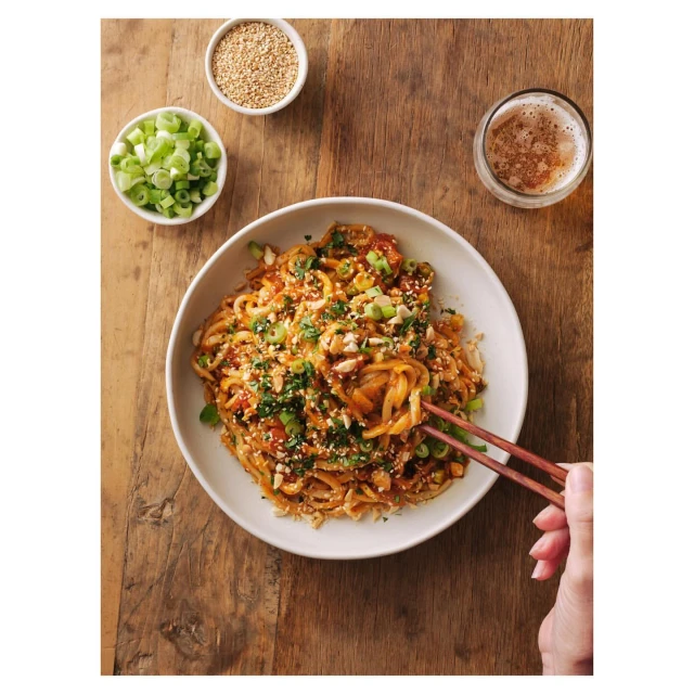 Overhead shot of noodles with garnishes and chopsticks presented on a wooden table next to a beer and bowls of sesame seeds and scallions.