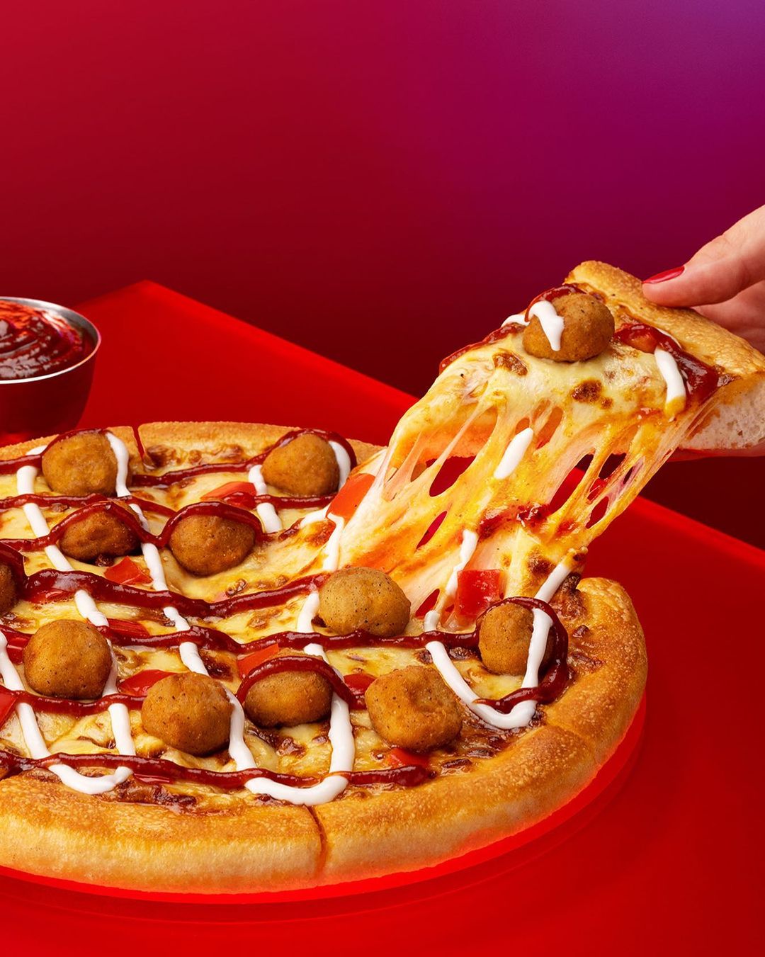 A slice of pizza is being held in a hand over a whole pizza with small meatballs and a red sauce drizzle, shot on a red surface.