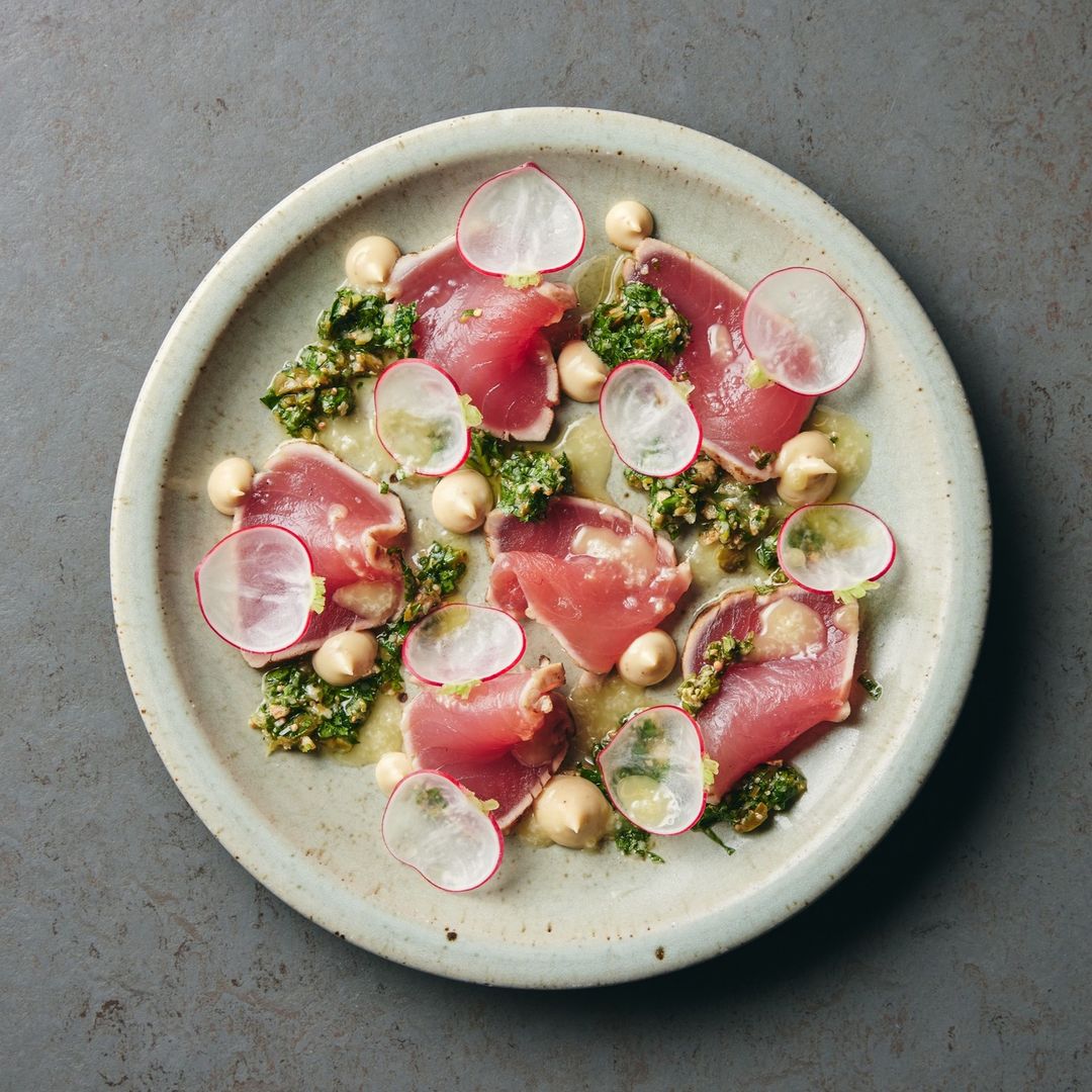 Overhead shot of seared tuna slices on a plate, garnished with radish, pesto, and cream dots on a gray surface.