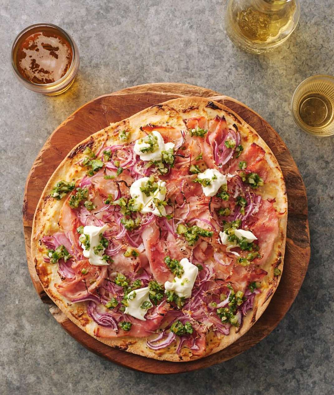 Overhead shot of pizza on a wooden board, topped with prosciutto, red onions, pesto, and dollops of ricotta cheese, with drinks in the background.