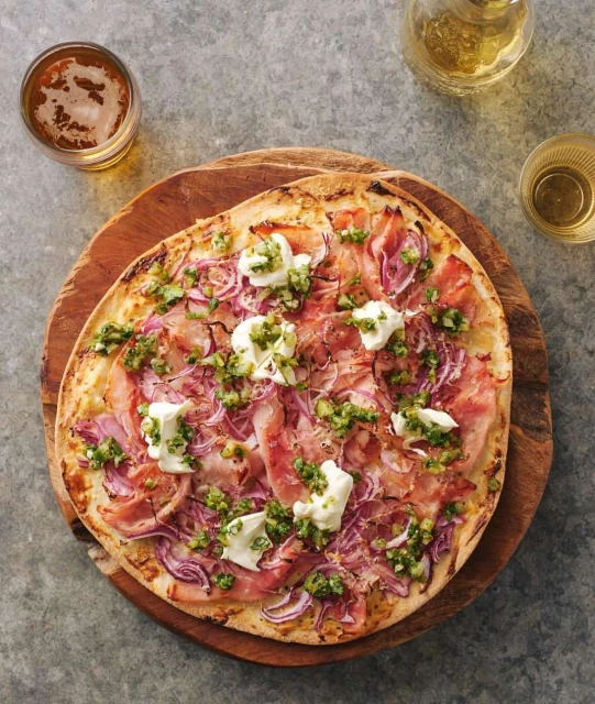 Overhead shot of pizza on a wooden board, topped with prosciutto, red onions, pesto, and dollops of ricotta cheese, with drinks in the background.