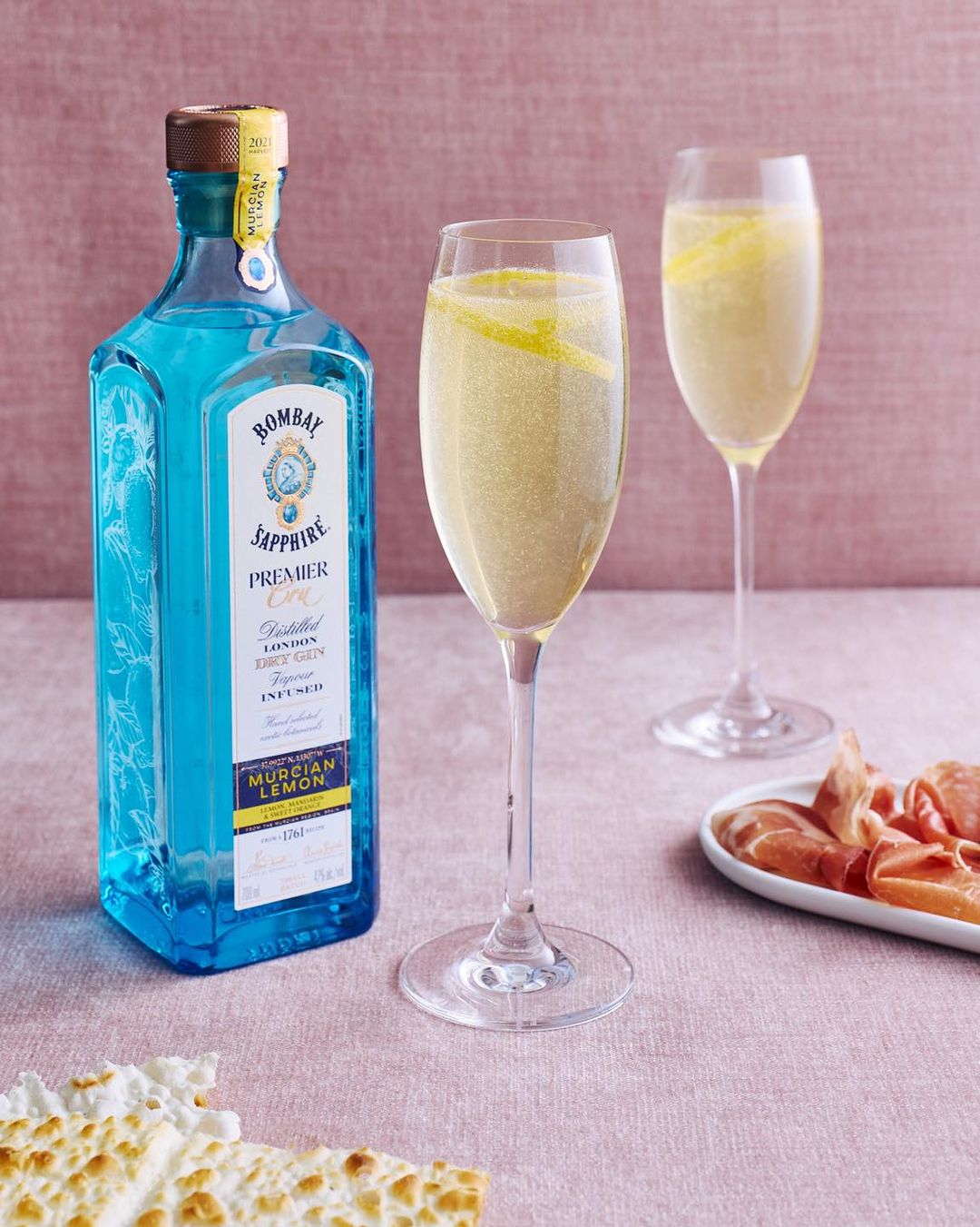 A bottle of Bombay Sapphire Premier Cru gin and two champagne flutes filled with sparkling drinks and lemon twists are on a pink table.