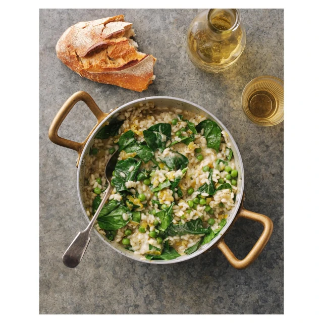 Overhead shot of a pot of risotto with spinach and peas, alongside bread and a bottle of oil on a grey textured surface.