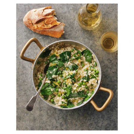 Overhead shot of a pot of risotto with spinach and peas, alongside bread and a bottle of oil on a grey textured surface.