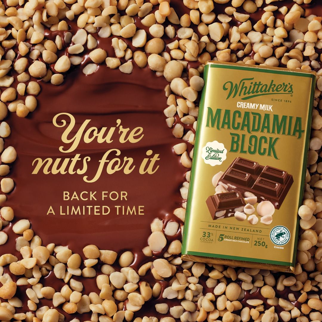 Whittaker's creamy milk chocolate bar with macadamia nuts surrounded by loose macadamia nuts on a brown reflective surface.
