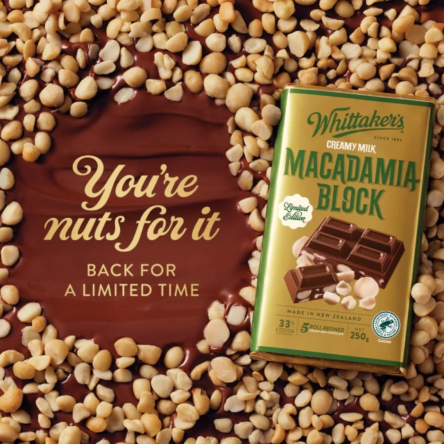 Whittaker's creamy milk chocolate bar with macadamia nuts surrounded by loose macadamia nuts on a brown reflective surface.