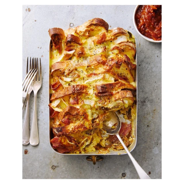 Overhead shot of a savoury strata bake in a metal baking pan, garnished with pepper and served with a side of red relish.