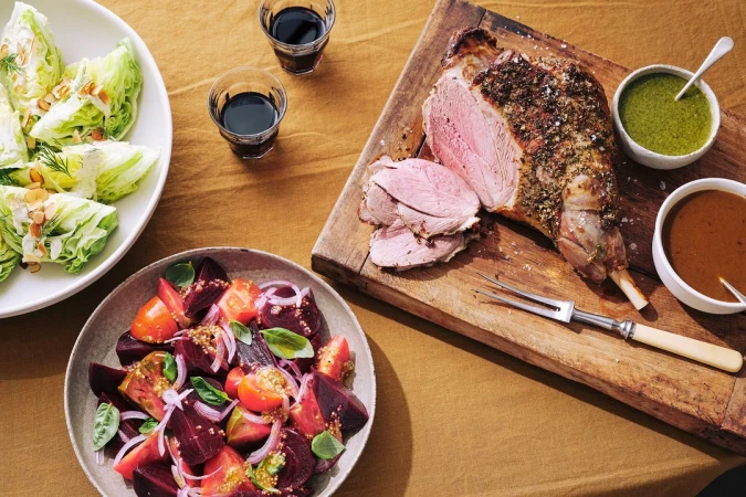 Overhead shot of roasted lamb leg on a wooden board with bowls of sauce, beet salad, lettuce salad, and glasses of dark liquid.