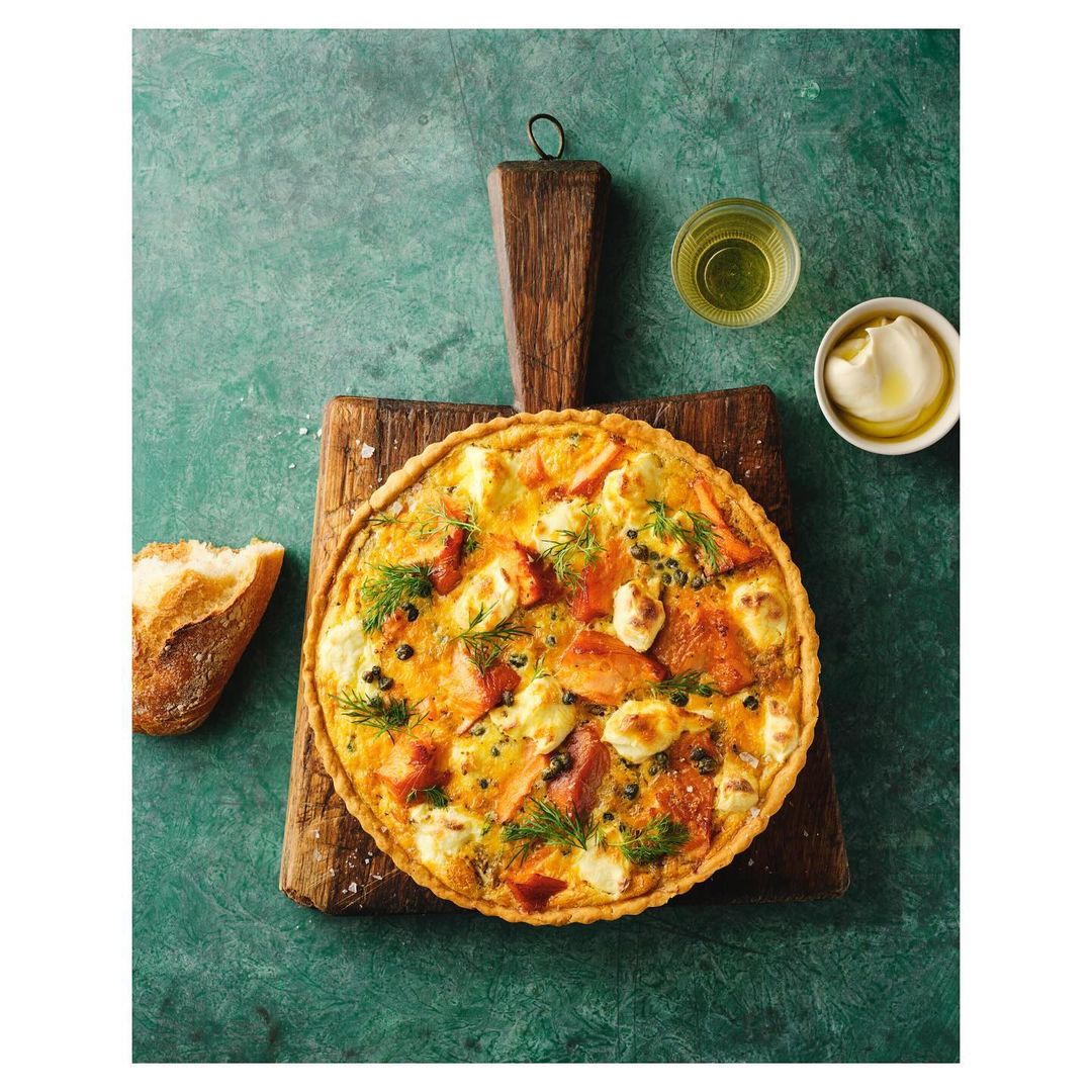 Overhead shot of salmon and cheese quiche on a wooden board with bread, oil, and dip against a textured green background.