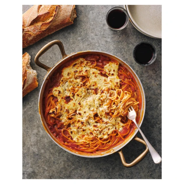 Overhead shot of spaghetti with tomato sauce and melted cheese in a copper pan, alongside bread and wine glasses on a gray surface.