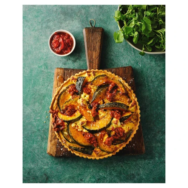 Overhead shot of a savory tart with squash and tomatoes on a wooden cutting board with a bowl of tomato sauce and greens.