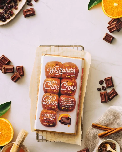 Overhead shot of Whittaker's Choc Cross Bun chocolate bar surrounded by loose chocolate pieces, orange slices, and baking rack on white marble.