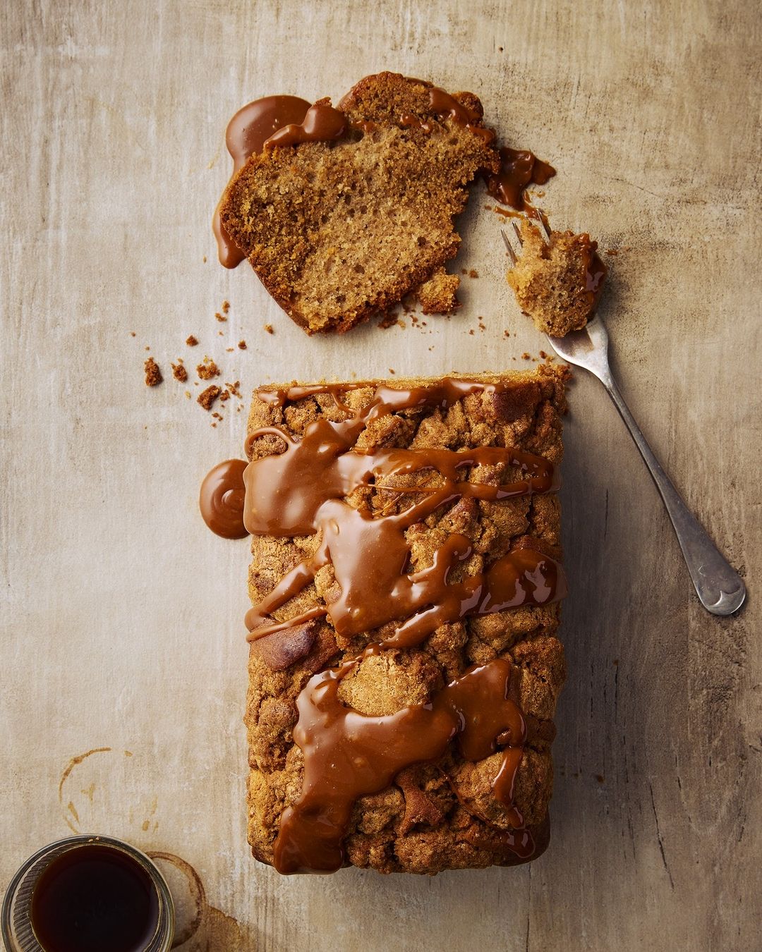 Overhead shot of a golden brown loaf cake with caramel sauce, alongside a slice on a fork and a coffee cup on a neutral surface.