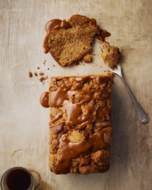 Overhead shot of a golden brown loaf cake with caramel sauce, alongside a slice on a fork and a coffee cup on a neutral surface.