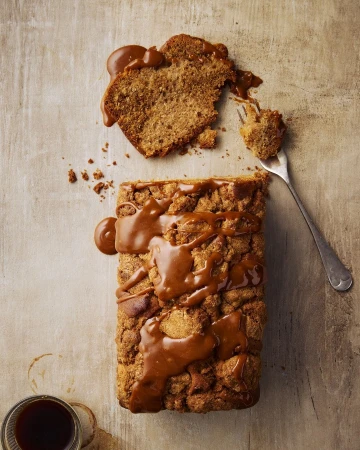 Overhead shot of a golden brown loaf cake with caramel sauce, alongside a slice on a fork and a coffee cup on a neutral surface.