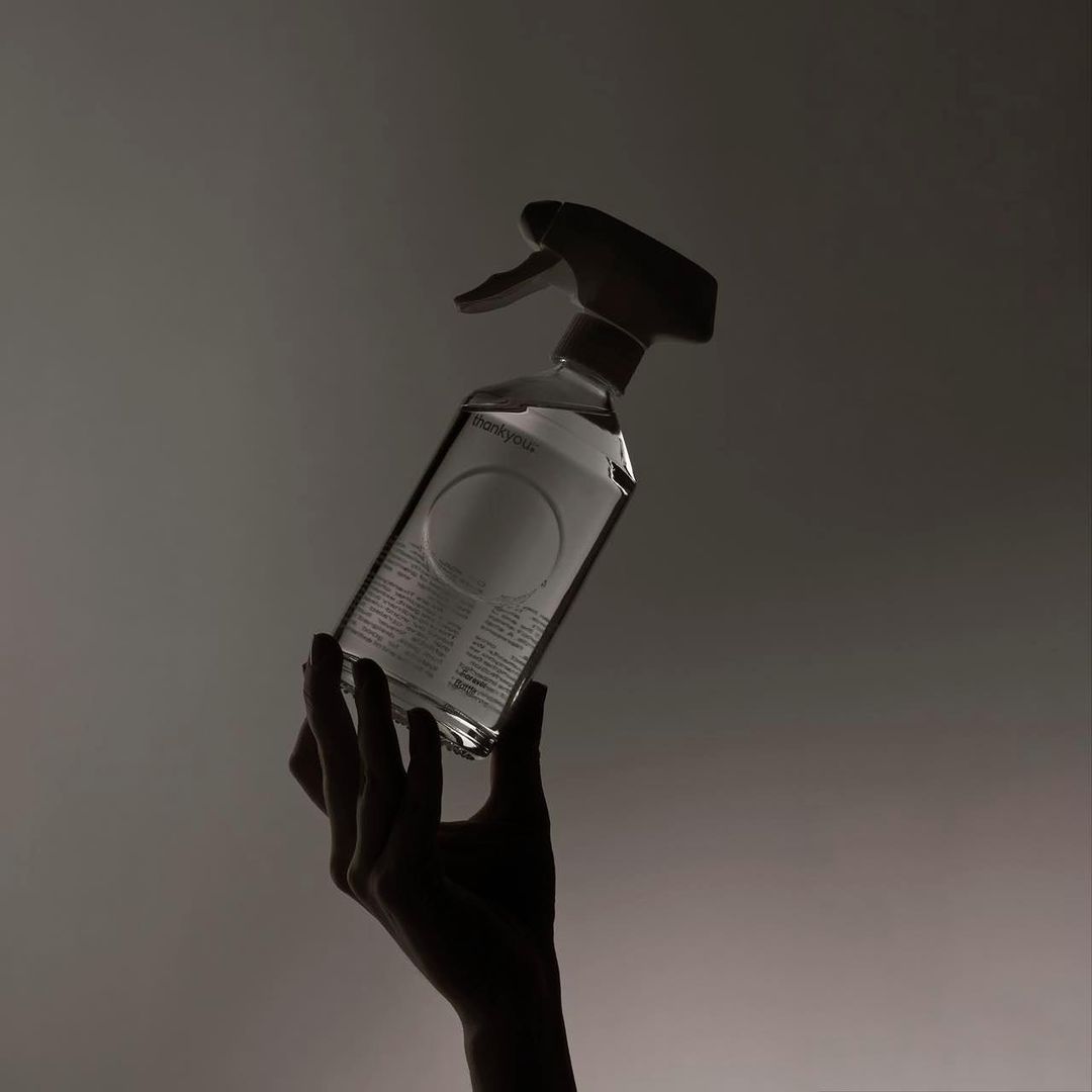 Close-up of a hand holding a clear spray bottle with a white label, backlit against a diffused gradient background, studio shot.