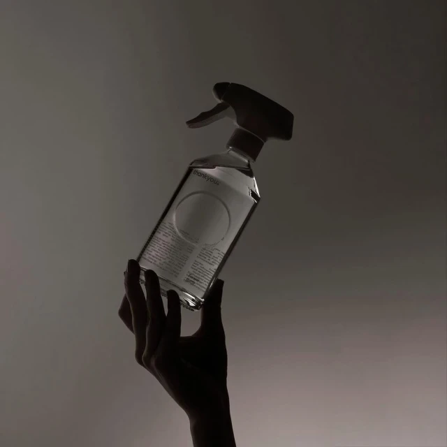 Close-up of a hand holding a clear spray bottle with a white label, backlit against a diffused gradient background, studio shot.