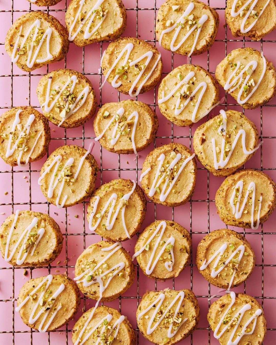 Overhead shot of dozens of round cookies decorated with white icing drizzle and chopped pistachios, arranged on a wire rack against pink.