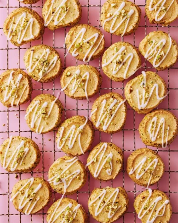 Overhead shot of dozens of round cookies decorated with white icing drizzle and chopped pistachios, arranged on a wire rack against pink.