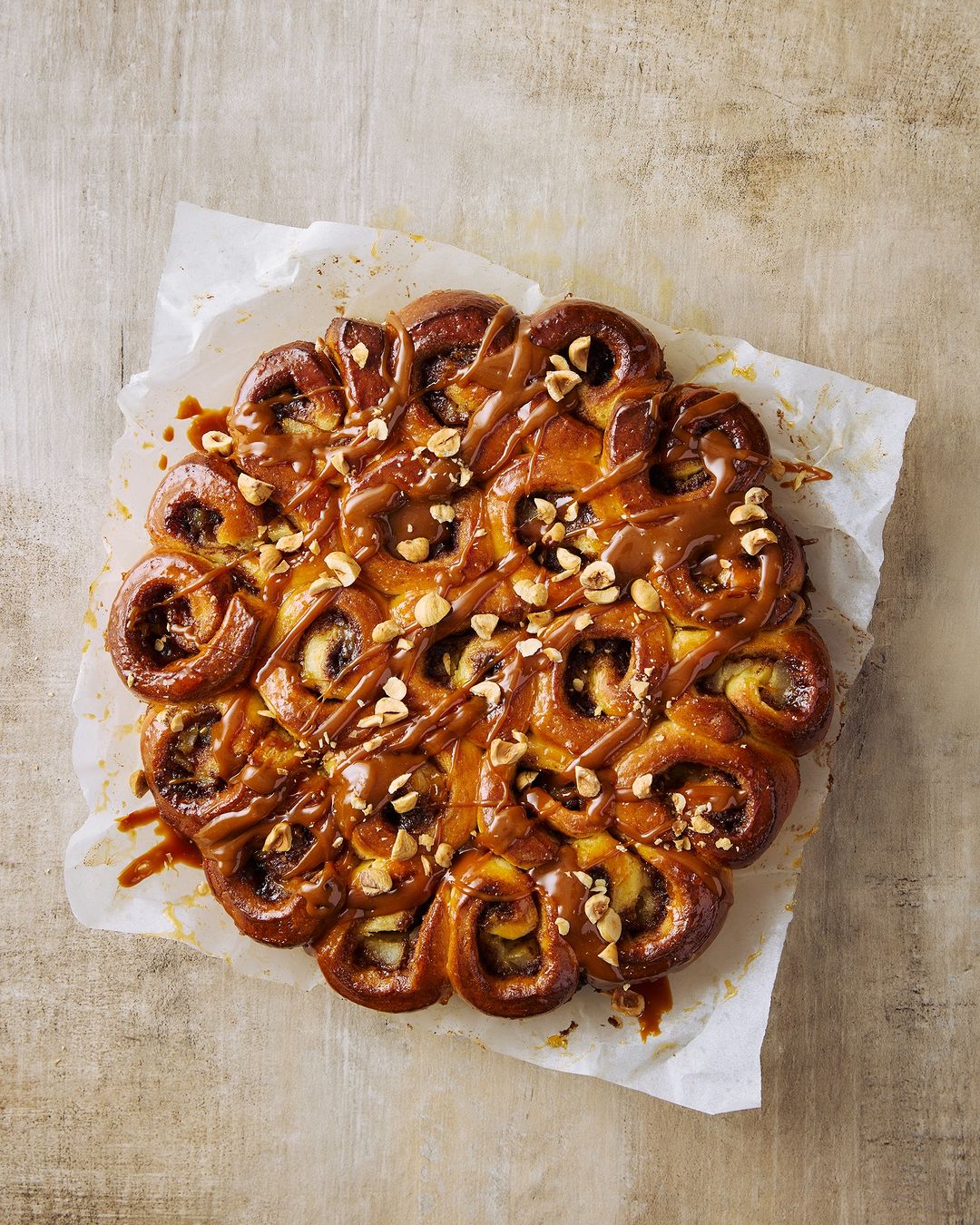 Overhead shot of a sweet pastry roll cake drizzled with caramel and topped with nuts on parchment paper with beige background.