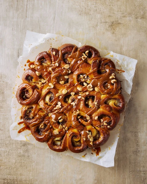 Overhead shot of a sweet pastry roll cake drizzled with caramel and topped with nuts on parchment paper with beige background.