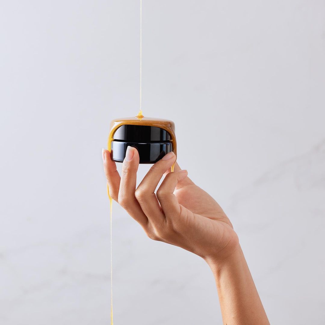 A woman's hand holds a dark jar with honey pouring over it, against a white background. Focus on hand and product.