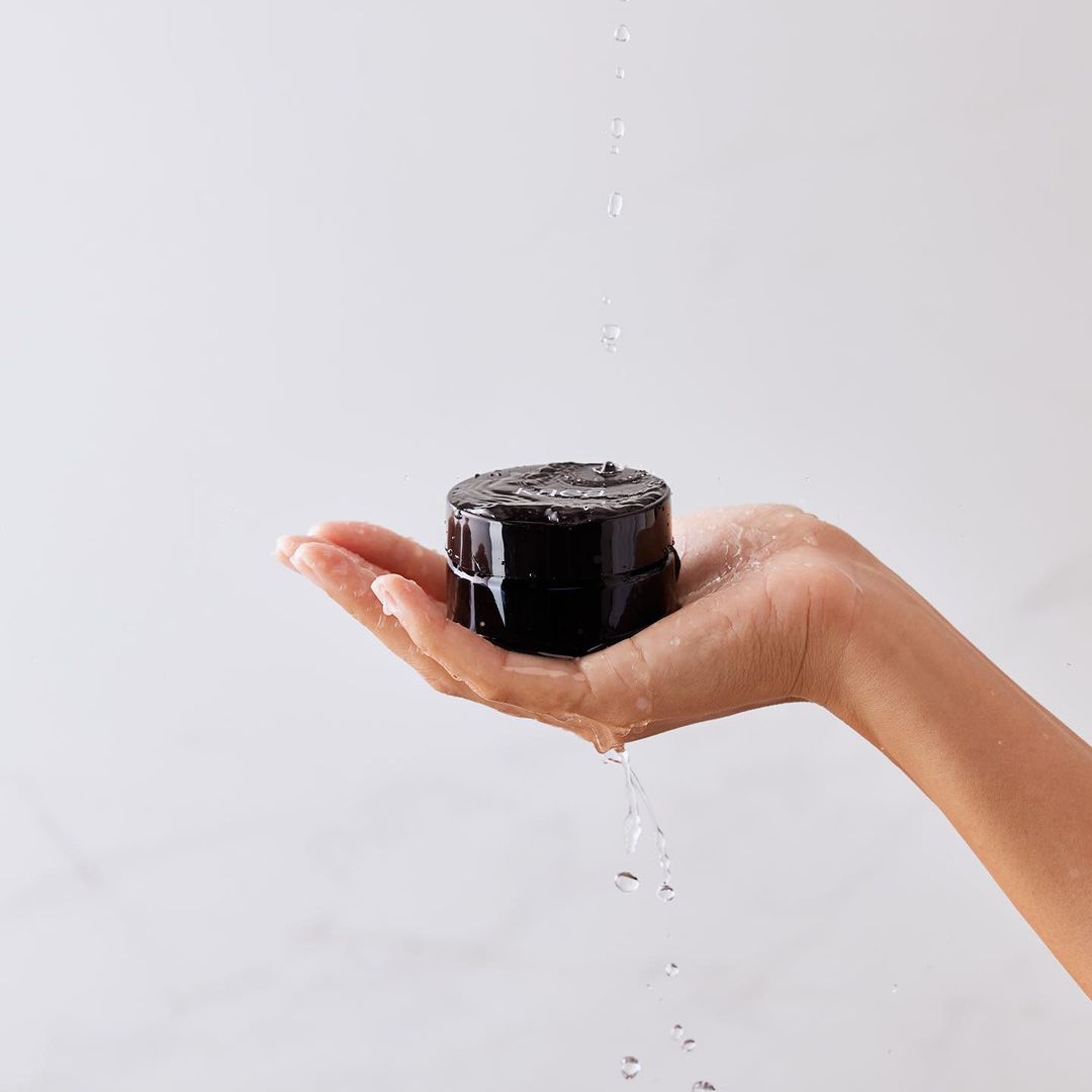 Close-up of a woman's hand holding a black cosmetic jar under streaming water against a clean, minimalist white background.
