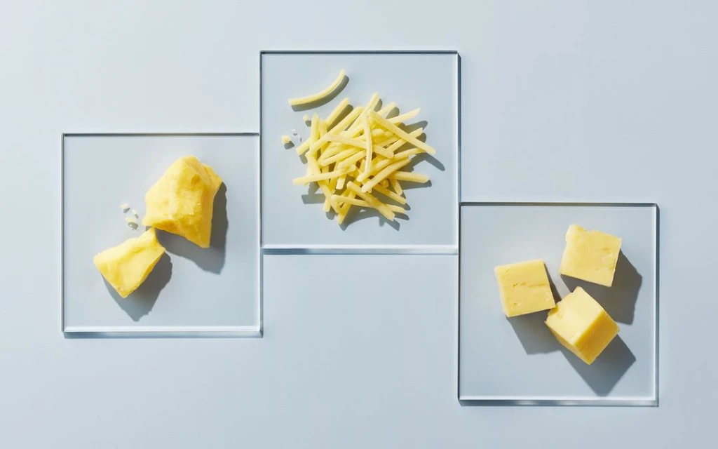 Three clear glass plates feature yellow cheese pieces cut into different shapes, all illuminated by a bright, clean studio light.