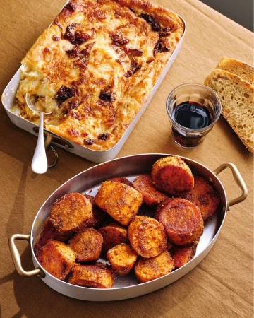 Overhead shot of two dishes: a casserole in a metal baking pan, served with roasted root vegetables, bread, and a glass of red wine.