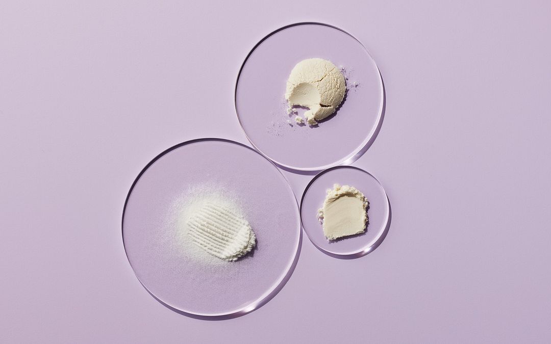 Overhead shot of skincare powders on clear acrylic discs against a soft purple backdrop, suitable for beauty and wellness product presentation.