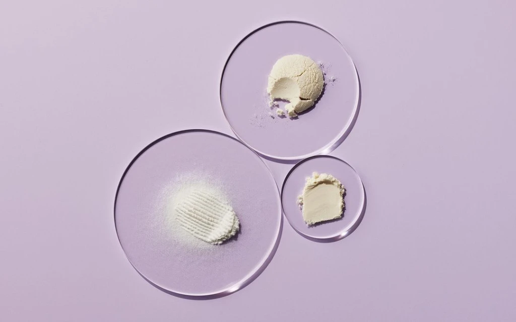 Overhead shot of skincare powders on clear acrylic discs against a soft purple backdrop, suitable for beauty and wellness product presentation.