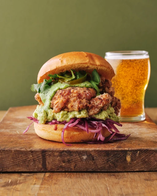 Fried chicken sandwich on a wooden board with a beer glass. Bun with shredded red cabbage and avocado sauce. Green background.