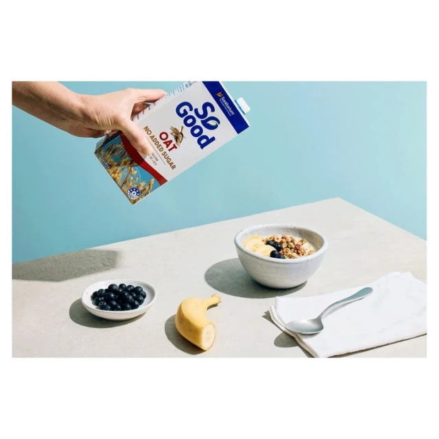 Person holding a carton of So Good oat milk next to a bowl of cereal, a side dish of blueberries, a banana, and a spoon on a light table.