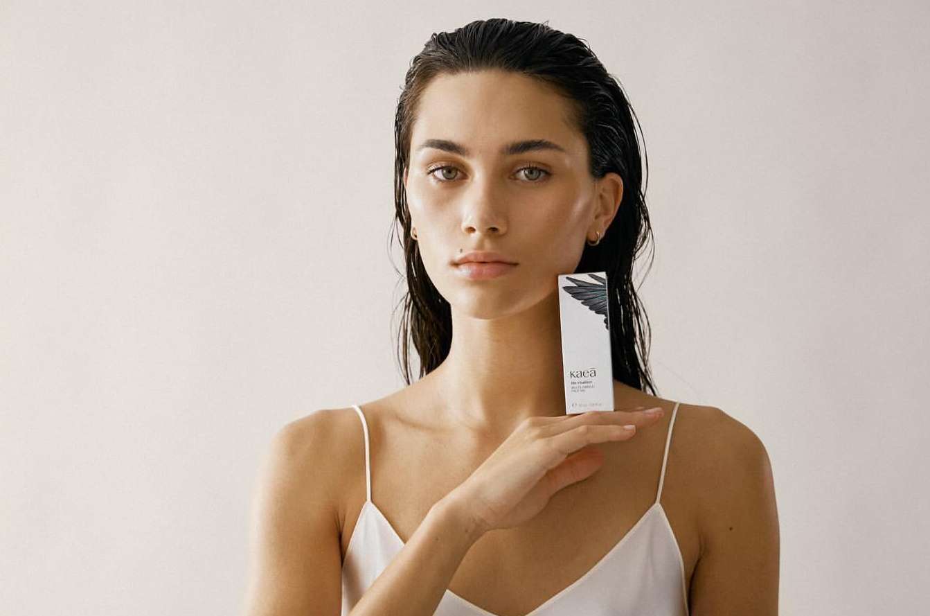 Adult woman with wet hair and white top holding a box of skincare product in a light-colored indoor setting. Beauty and fashion.