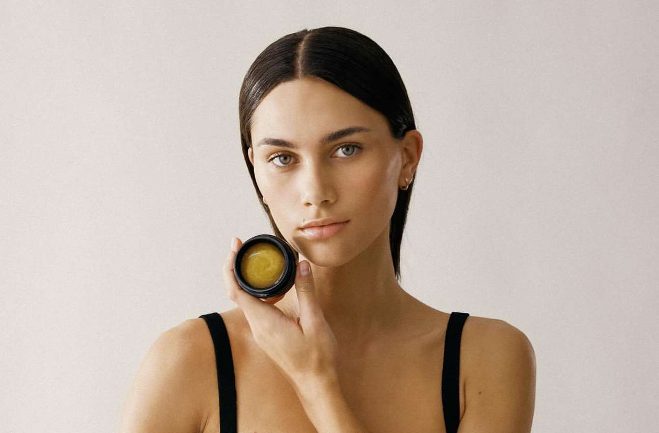 Close-up shot of a woman holding a jar of beauty product. The woman has long, dark hair and a neutral expression. Cream background.