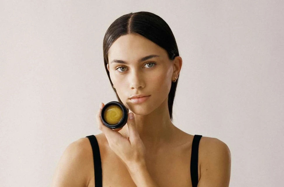 Close-up shot of a woman holding a jar of beauty product. The woman has long, dark hair and a neutral expression. Cream background.