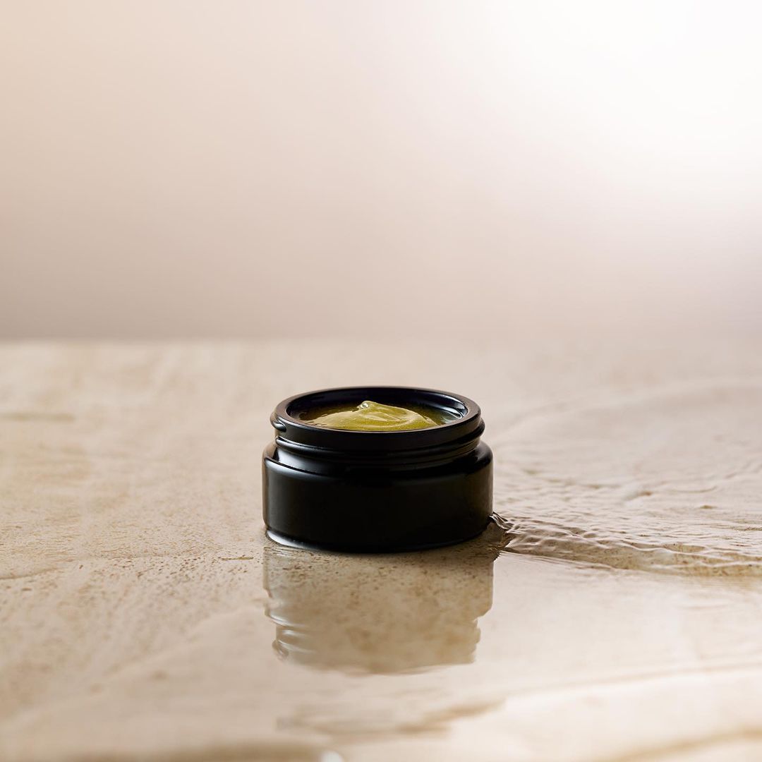 A black jar of skin cream sits on a textured surface with a neutral background. The container reflects light, emphasizing the product.