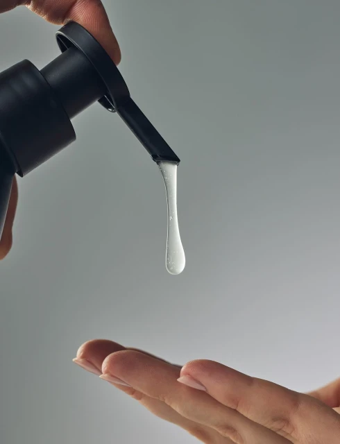 Close-up of a clear drop of liquid dispensing from a black pump dispenser onto the outstretched fingers of an adult female hand.