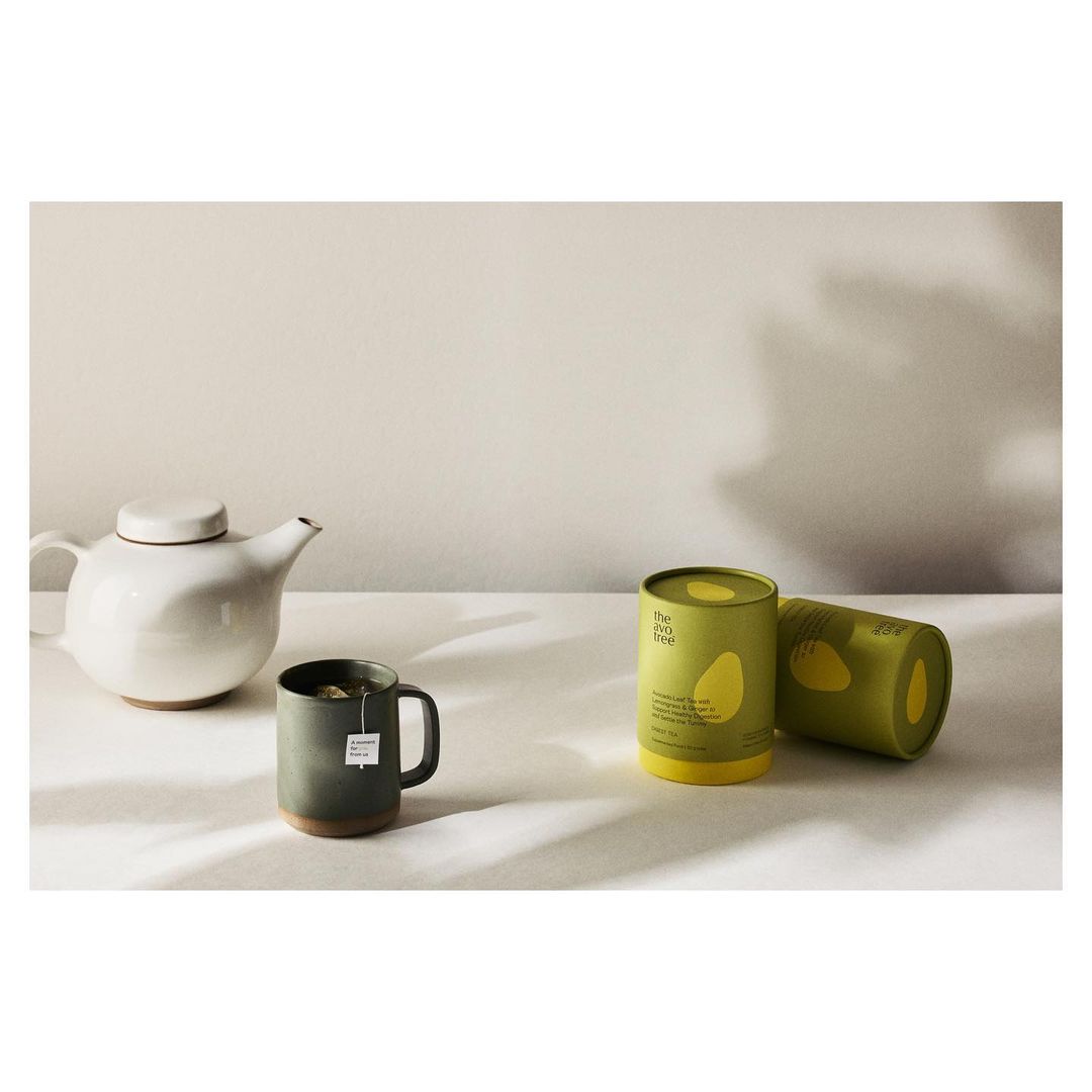 Still life featuring a white teapot, a green mug with a teabag, and two green and yellow tea canisters on a white surface.