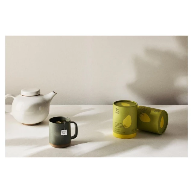 Still life featuring a white teapot, a green mug with a teabag, and two green and yellow tea canisters on a white surface.