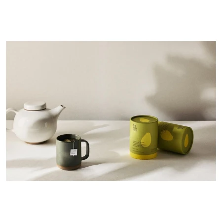 Still life featuring a white teapot, a green mug with a teabag, and two green and yellow tea canisters on a white surface.