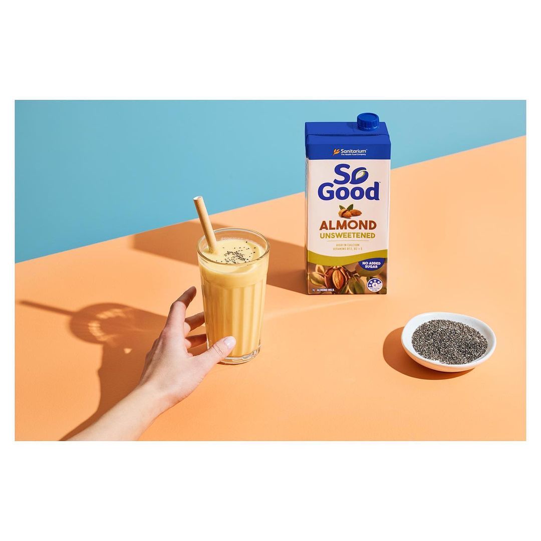 Almond milk smoothie in a glass with a straw, next to a carton of So Good almond milk and a bowl of chia seeds on a table.