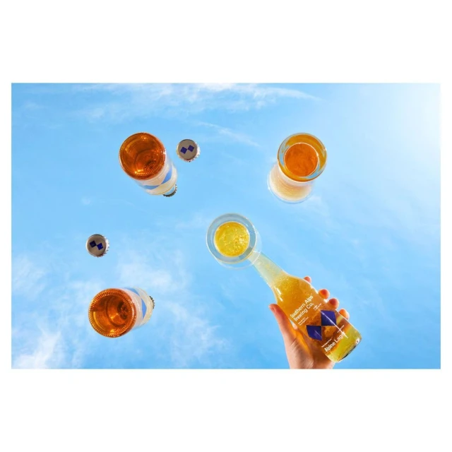 Low-angle shot of a hand holding a bottle of citrus drink with open bottles and caps floating mid-air against a blue sky with clouds.