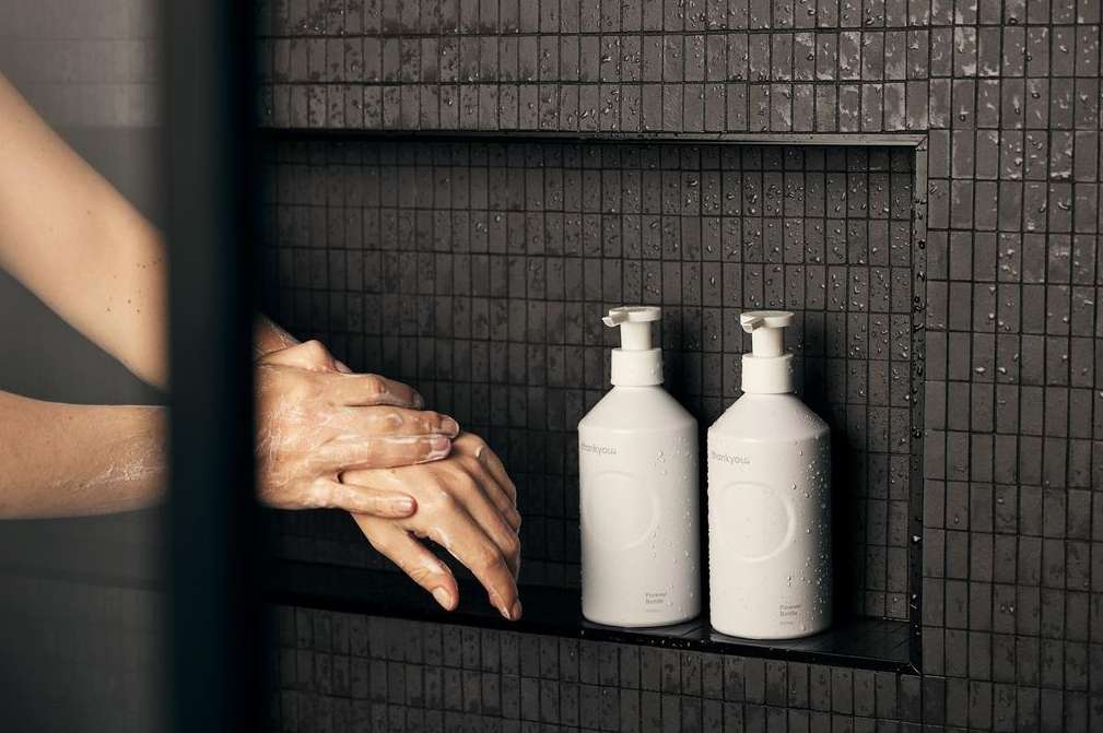Adult washing hands in a shower with a pair of white pump bottles on a tiled shelf, promoting cleanliness and self-care.