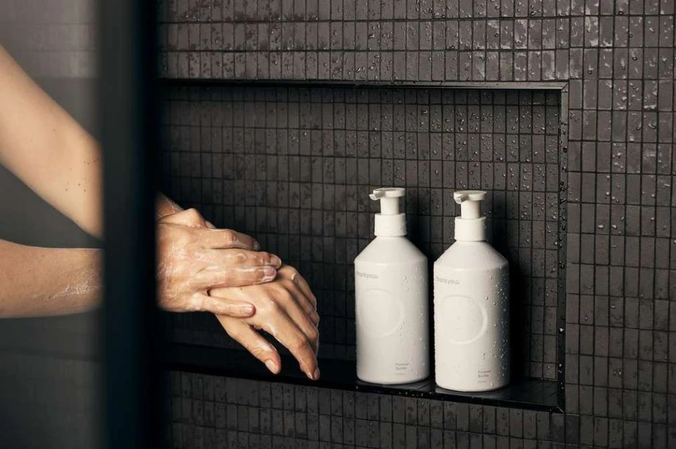Adult washing hands in a shower with a pair of white pump bottles on a tiled shelf, promoting cleanliness and self-care.