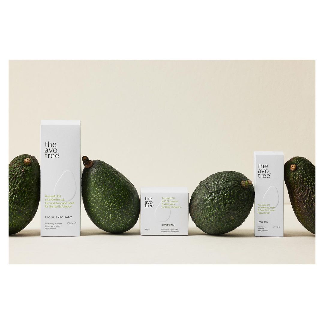 Close-up product shot of The Avo Tree skincare line featuring facial exfoliant, day cream, and face oil, accented by fresh avocados.