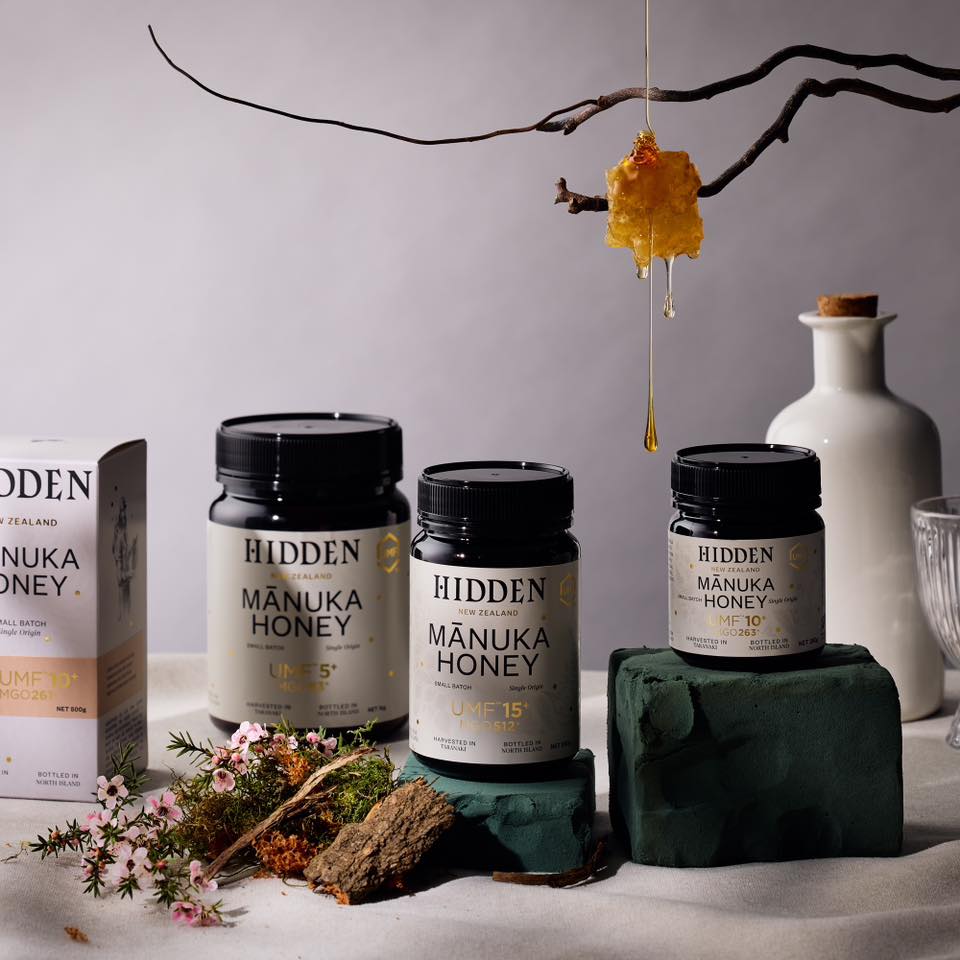 Still life shot of Hidden New Zealand Manuka Honey product line in jars and box, next to floral and branch decorations on a table.